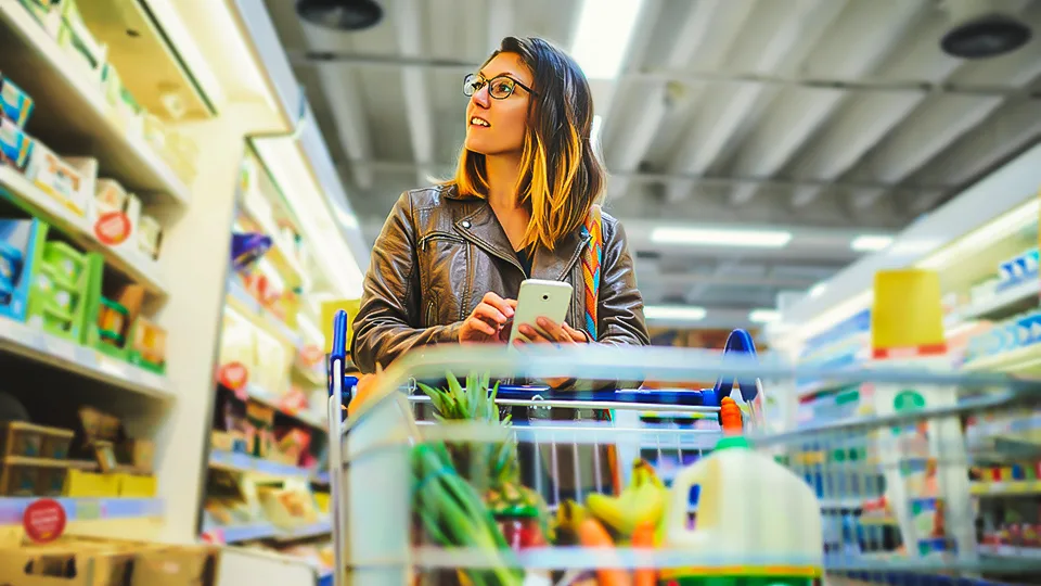 A woman shops in a grocery store aisle with a cart filled with fresh produce and locally sourced items.