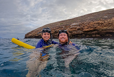 Two guests floating together near Molokini Crater, smiling with a flotation noodle and masks pushed up.