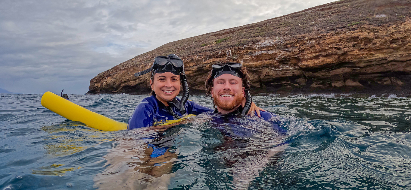 Two guests floating together near Molokini Crater, smiling with a flotation noodle and masks pushed up.